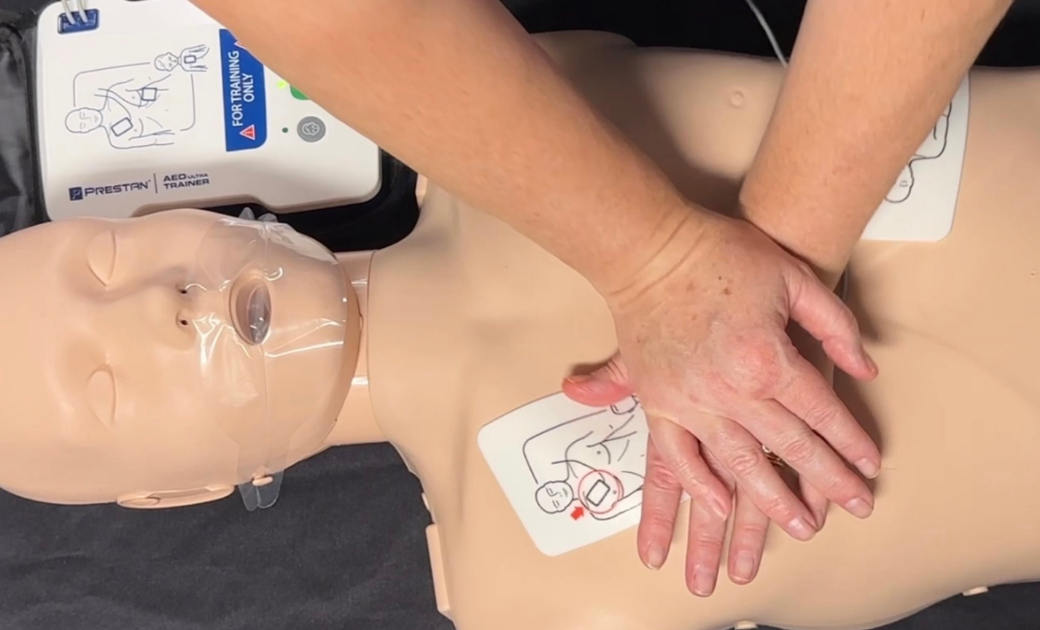 A person performing CPR on a training mannequin, with hands positioned over the chest. Two AED pads are visible on the mannequin, along with an automated external defibrillator nearby.