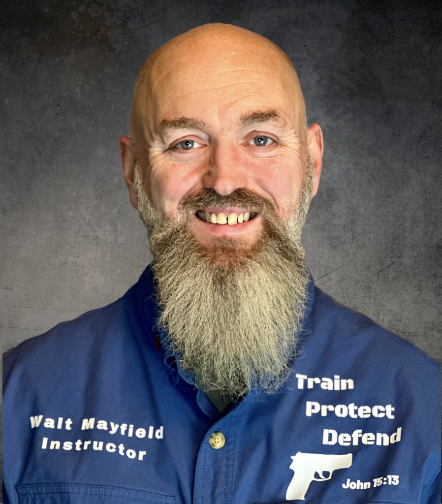 A smiling man with a long beard wearing a blue shirt that reads "Train Protect Defend" and "Walt Mayfield Instructor" in white text, with a dark, textured background.