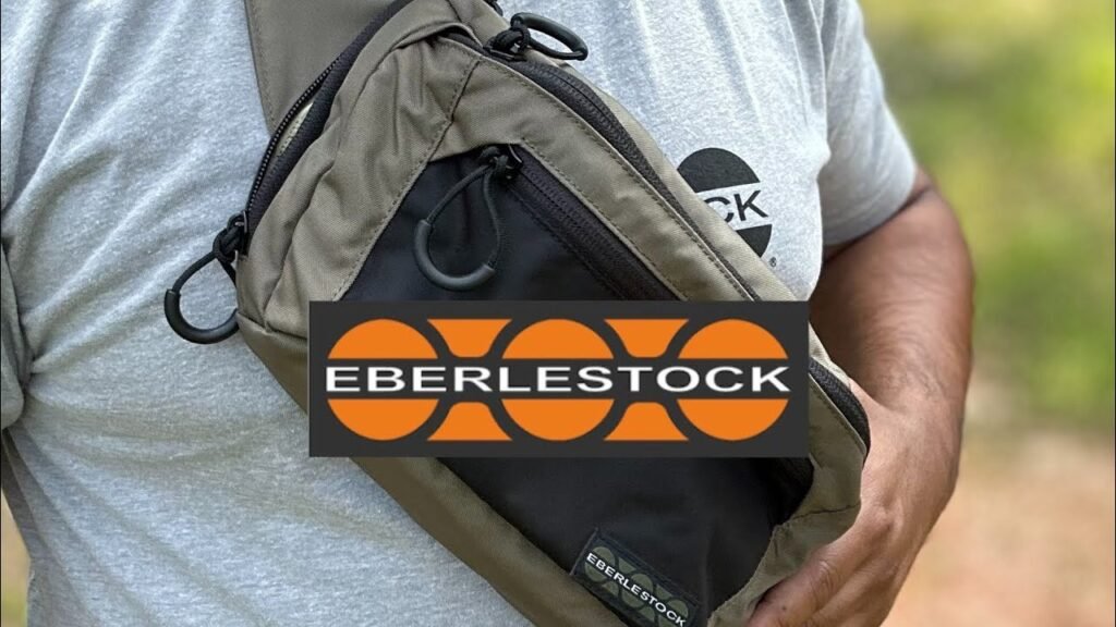 A person wearing a light gray t-shirt holds a tan and black Eberlestock sling bag. The bag is positioned across the body, showing its design and branding.